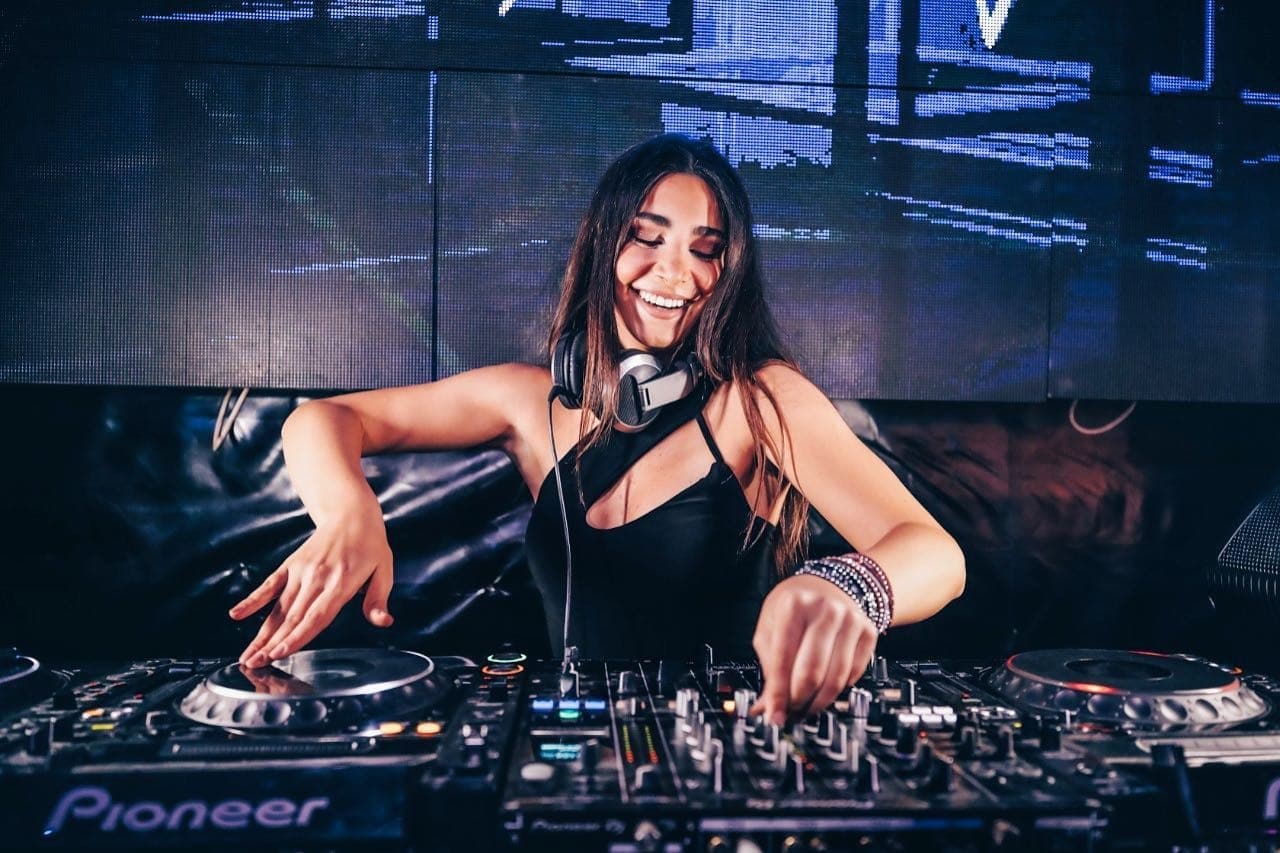 Lebanese DJ Chloe Kteily mixing on CDJ players behind a lit DJ booth, hands on the decks during a live set