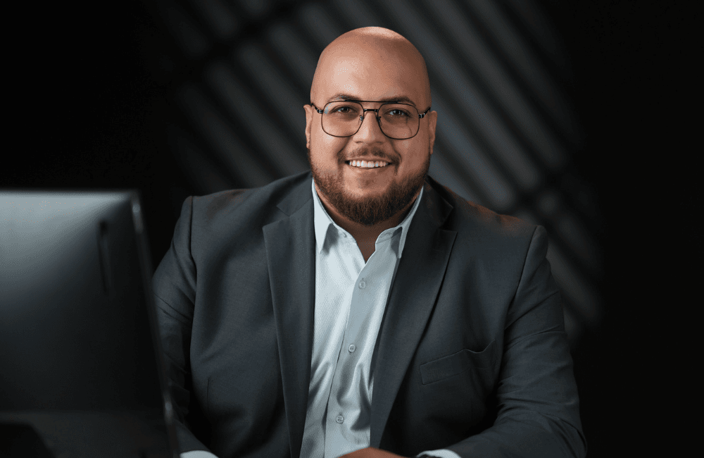 Ali Haddad, Media Specialist at ZXCM, smiling in a dark suit and light shirt seated at his desk against a dark studio background.