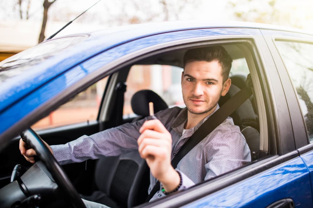 A young man sits in the driver's seat of a blue car, seatbelt on, holding up a car key while resting his other hand on the steering wheel.