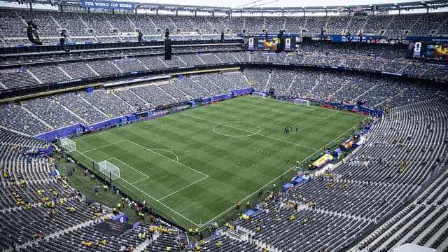Wide aerial view of a packed World Cup stadium with a bright green natural-grass pitch, players warming up on the field, and FIFA World Cup 2026 branding on the scoreboards.