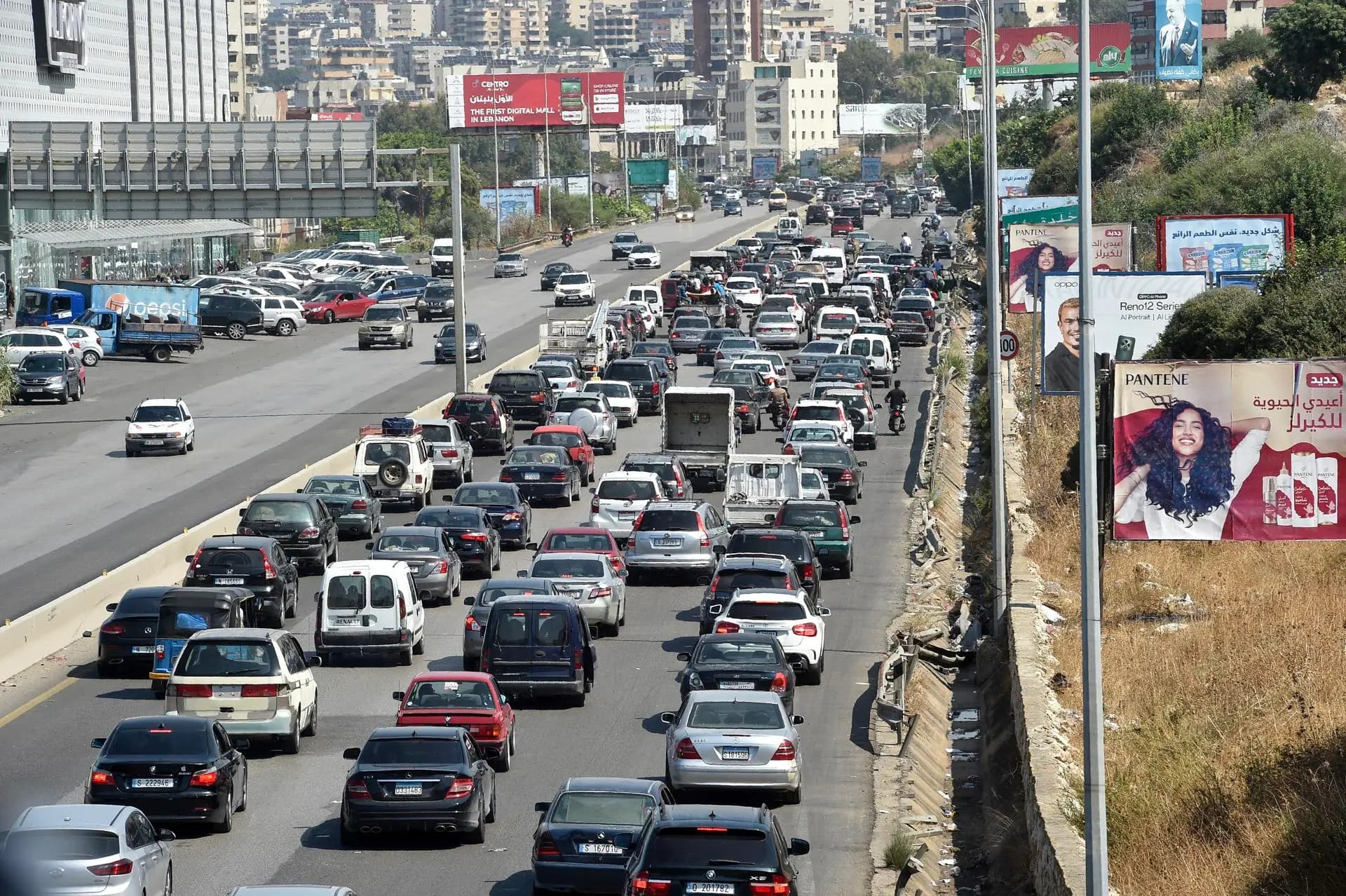 Heavy weekend traffic backed up on a multi-lane highway leading into Beirut, with cars and vans packed bumper to bumper, city buildings and billboards in the background.