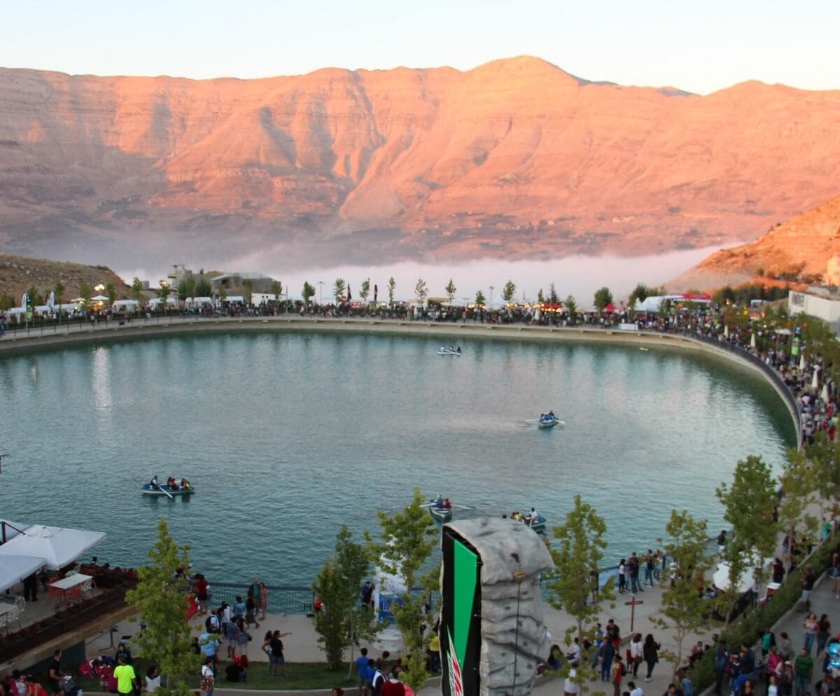 Crowds fill the promenade around Zaarour Club's oval mountain lake at sunset, with pedal boats on the water, a climbing wall and food stalls in the foreground, and pink-lit peaks and low fog behind.