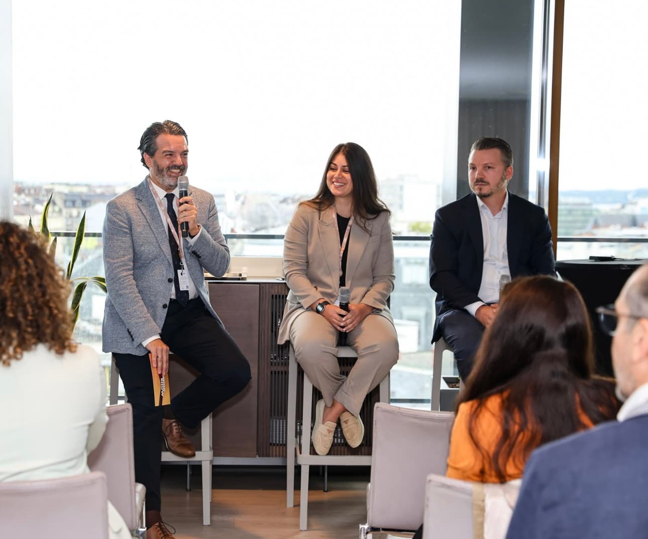 Sandra Helou, in a beige suit, sits between two male panelists in front of floor-to-ceiling windows overlooking a city skyline; the man on her right speaks into a microphone while she smiles, with an audience in the foreground.