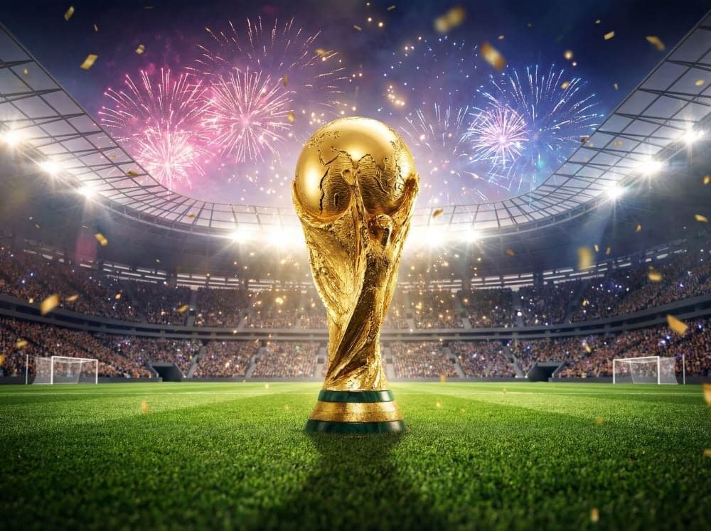 The golden FIFA World Cup trophy standing on a green stadium pitch, with fireworks and gold confetti lighting up a packed stadium at night