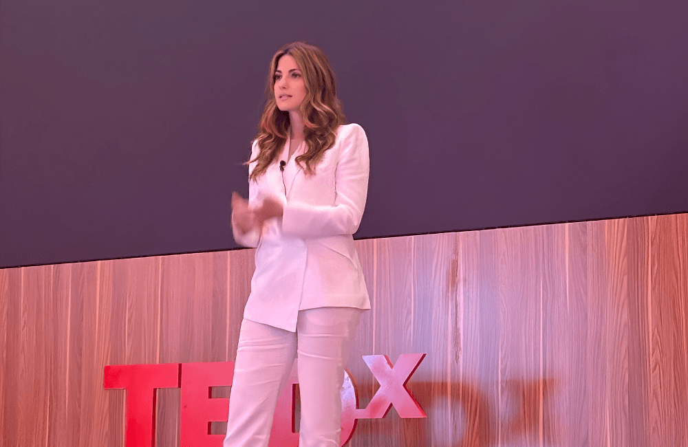 Christiane Nasr, founder of The Bowery Company, speaking on stage in a white suit at a TEDx event, mid-gesture, with red TEDx lettering on a wooden backdrop behind her.