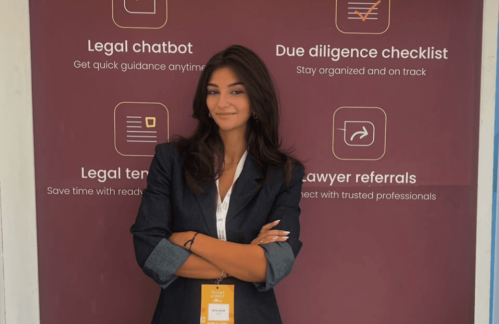 askLex founder May El Hachem stands with her arms crossed in front of a maroon askLex banner that lists the app's four features: legal chatbot, due diligence checklist, legal templates, and lawyer referrals. She wears a navy blazer and a summit speaker badge on a lanyard.