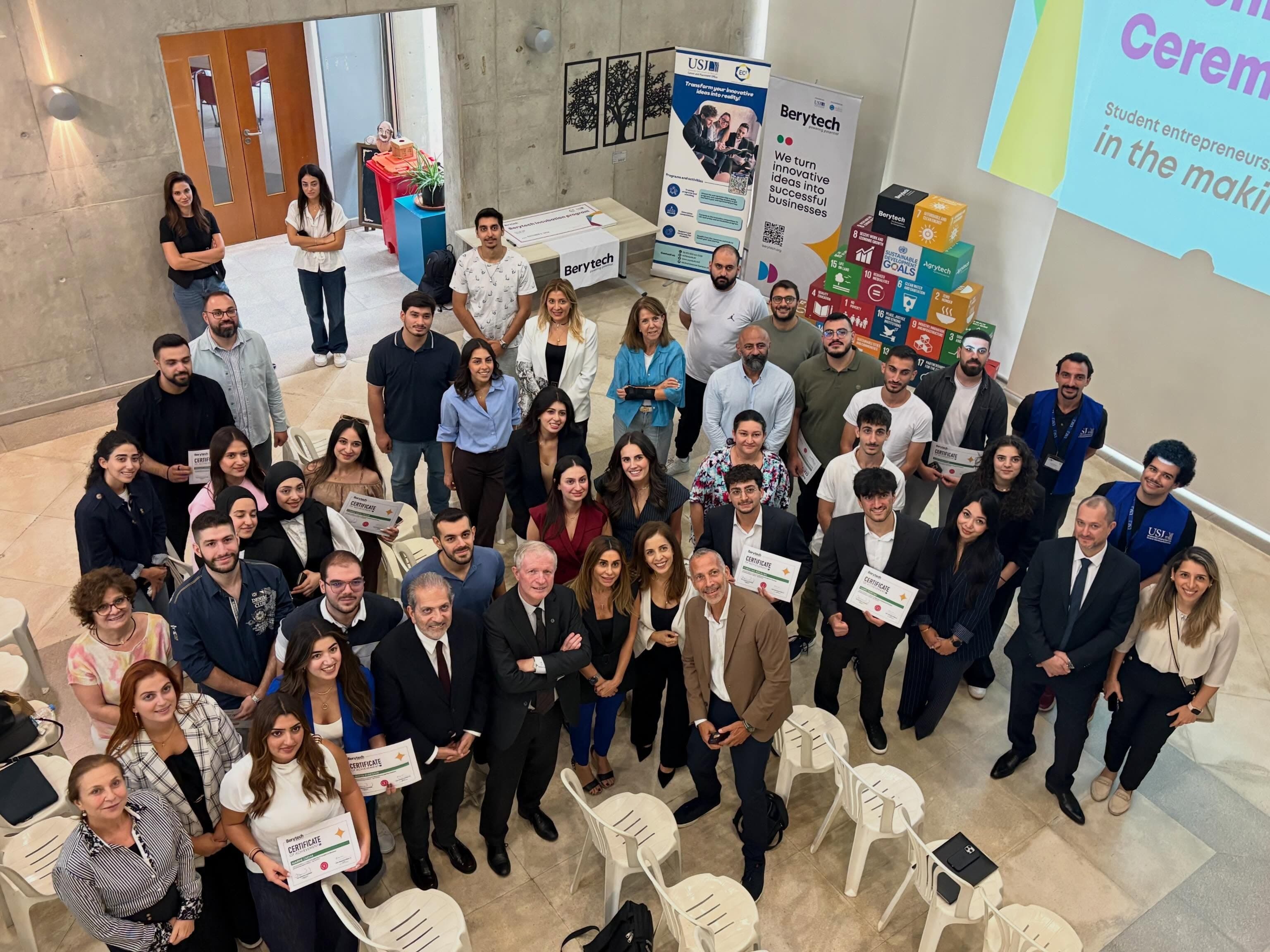 Overhead group photo of student entrepreneurs, mentors and organizers at the Berytech x USJ EC² Incubation Pitching Ceremony, with several founders holding Berytech certificates of achievement in front of Berytech and USJ banners and a stack of Sustainable Development Goals cubes