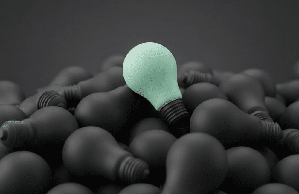 A single mint green lightbulb resting on top of a large pile of dark charcoal lightbulbs against a dark grey background.