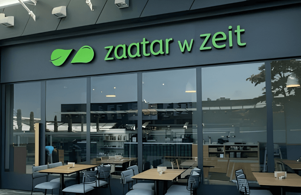 Zaatar w Zeit restaurant storefront with the green leaf logo and brand name on a dark grey facade, floor to ceiling windows, and wooden tables with grey chairs on the outdoor terrace